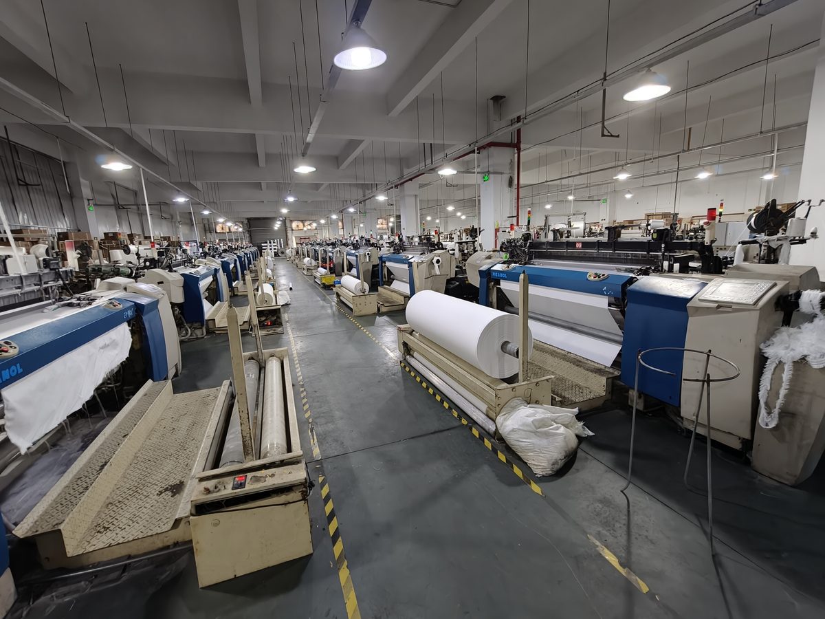 Rows of air-jet looms with white greige fabric rolls in the weaving shed